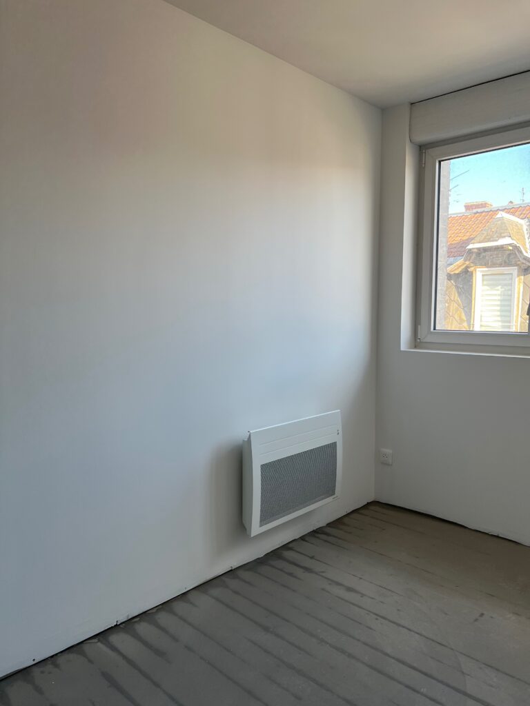 Chambre rénovée avec radiateur électrique mural et fenêtre.